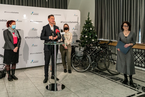 Cztery osoby na konferencji prasowej, w tle biała ścianka z logotypami Pomorze Zachodnie, choinka, a przy niej ustawione dwa rowery.