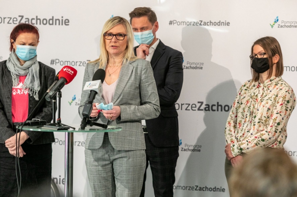 Anna Bańkowska, członkini zarządu województwa, podczas konferencji prasowej stoi przy stoliku z mikrofonami, za nią - marszałek Olgierd Geblewicz.