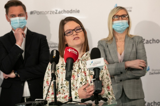 Kobieta stoi przy stoliku z radiowymi mikrofonami; za nią - kobieta i mężczyzna również uczestniczący w konferencji prasowej.